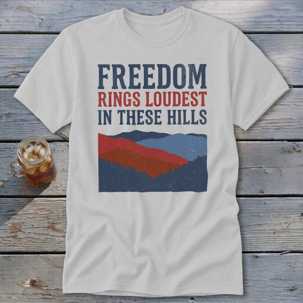 A light gray t-shirt with blue and red text reading Freedom Rings Loudest in These Hills above layered mountain graphics.