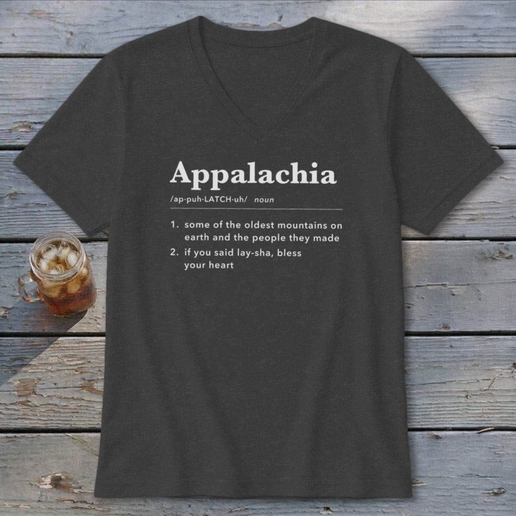 A dark gray V-neck T-shirt with white text defining the word Appalachia rests on a wooden surface beside a small glass of iced tea.