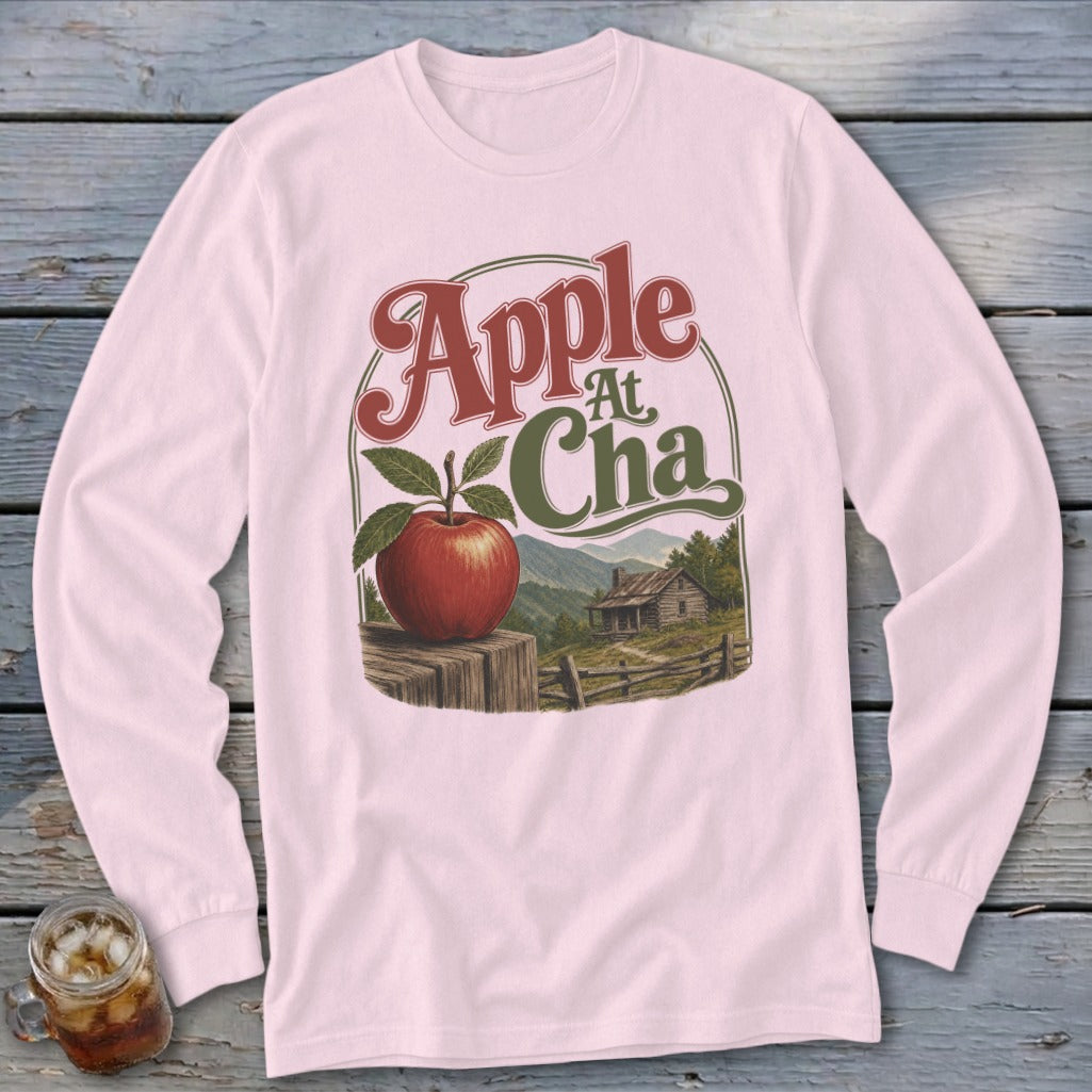 A light pink long-sleeve shirt featuring a retro graphic of an apple, a cabin, and mountains.