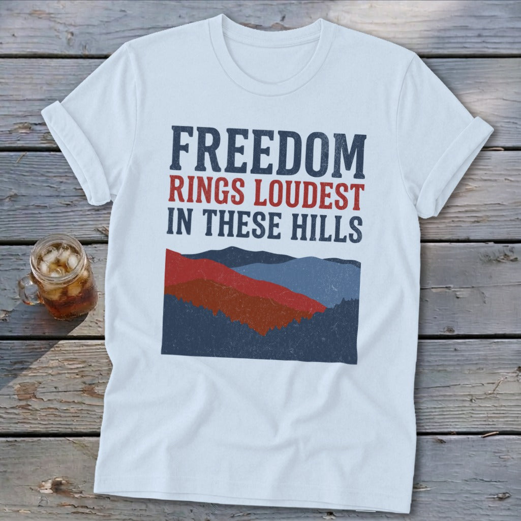 A light blue t-shirt with rolled sleeves rests on a weathered wooden surface, featuring text and mountain graphics.