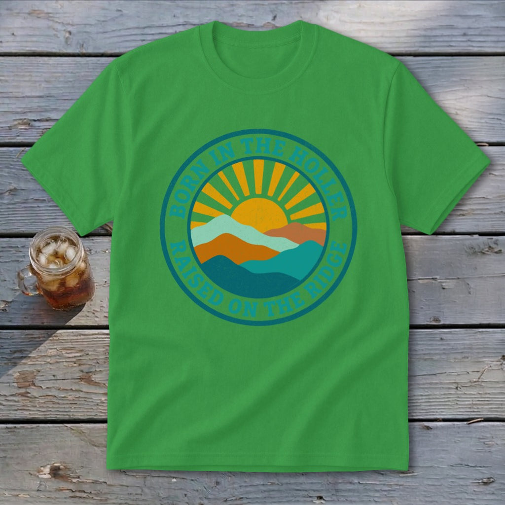 A green t-shirt featuring a circular graphic with mountains, a rising sun, and text that reads BORN IN THE HOLLER RAISED ON THE RIDGE.