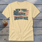 Cream t-shirt featuring a distressed graphic of the American flag and the text NEW YORK RAISED AMERICAN MADE.