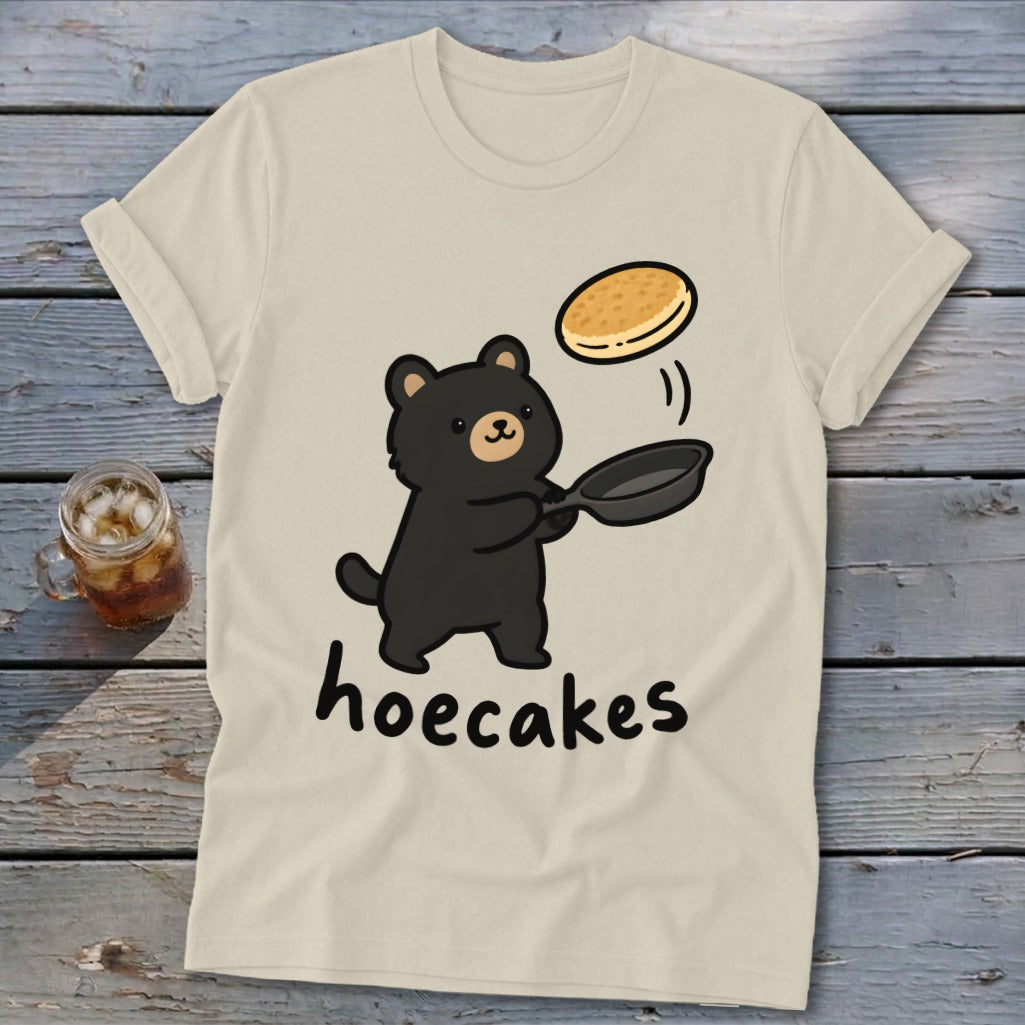 A cream-colored t-shirt displays an illustration of a black bear flipping a pancake with the text hoecakes.