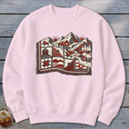A light pink crewneck sweatshirt featuring a graphic of a patchwork quilt merging into a mountain landscape with suns and trees.