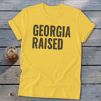 Yellow t-shirt with "GEORGIA RAISED" text, shown on a wooden surface.