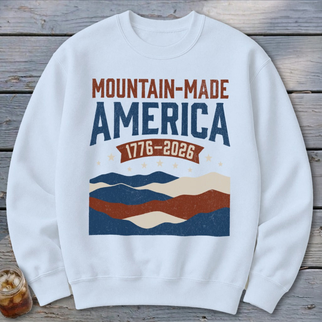 White crewneck sweatshirt featuring a distressed graphic with the text Mountain-Made America 1776-2026 over wavy patriotic stripes.
