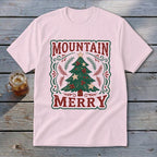 A light pink t-shirt featuring a vintage Christmas tree design with the text Mountain Merry on a wooden background.