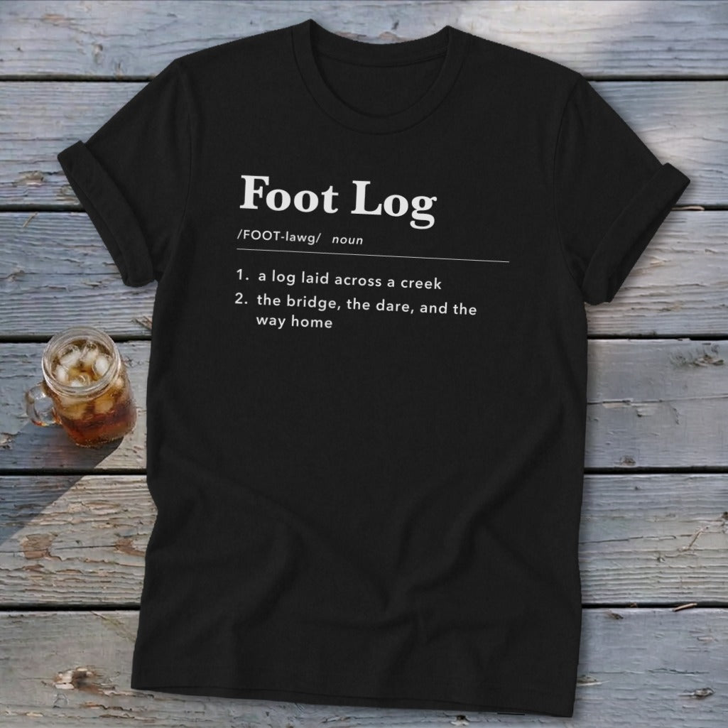 A black t-shirt printed with a dictionary definition of Foot Log on a rustic wooden background.