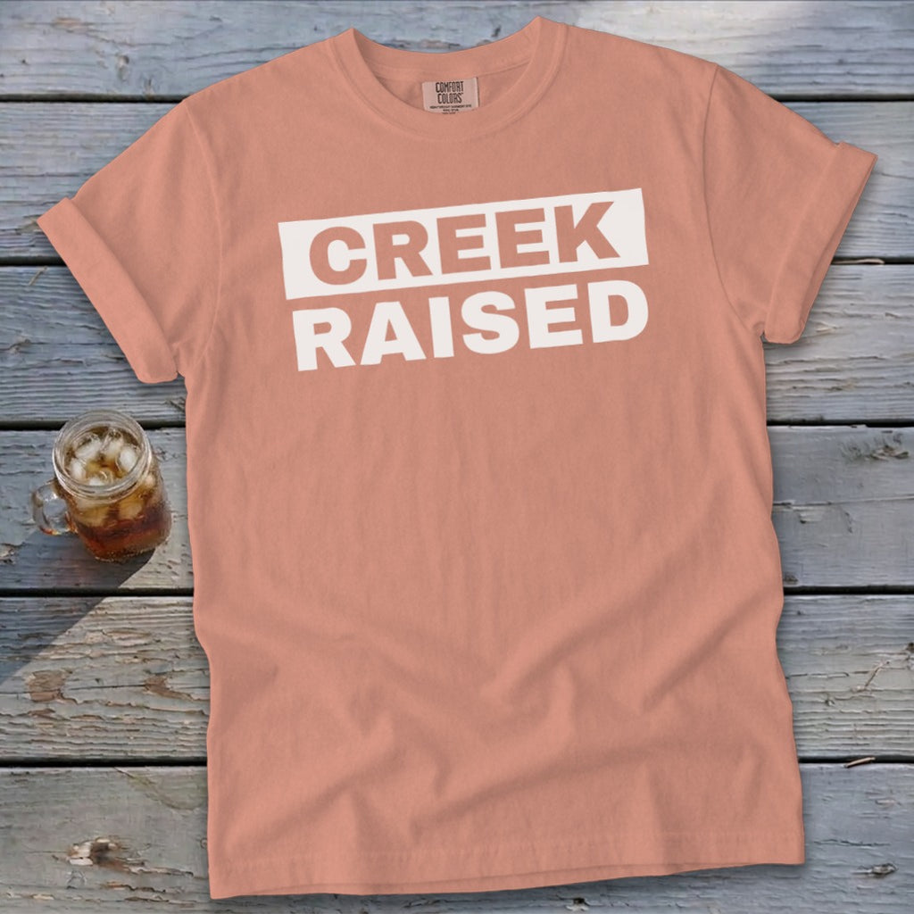 Peach t-shirt with white text "CREEK RAISED" and a glass of iced tea.
