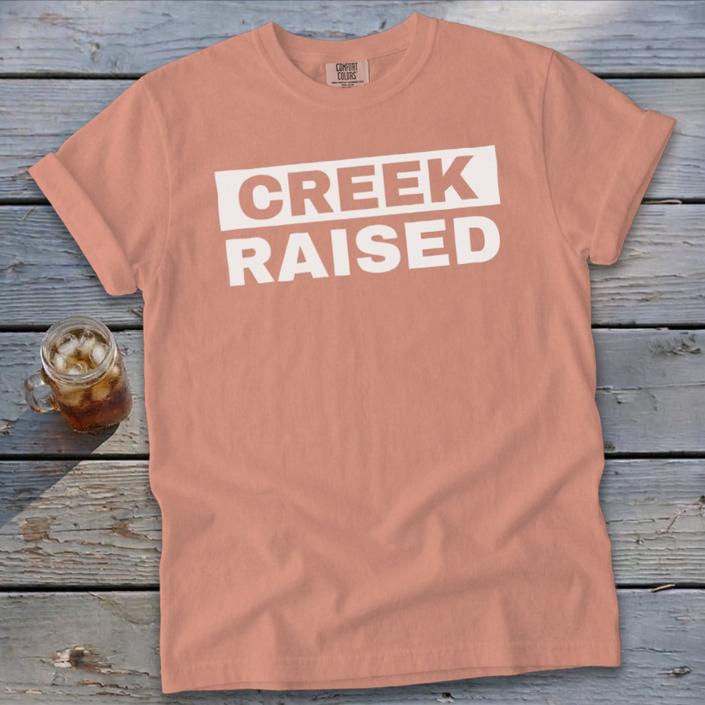Peach t-shirt with white text "CREEK RAISED" and a glass of iced tea.