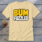 Cream crew-neck t-shirt featuring the text BUM FUZZLED in bold yellow and white letters, laid on a weathered wooden surface next to an iced beverage.