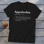 A black t-shirt printed with the dictionary definition of Appalachia lies flat on a rustic wooden table next to a glass of iced tea.
