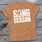 A camel-colored t-shirt with rolled sleeves displaying distressed white text reading SANG SEASON across the chest.