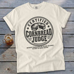 Off-white t-shirt featuring a circular graphic with text reading Certified Cornbread Judge and a sliced cornbread illustration.