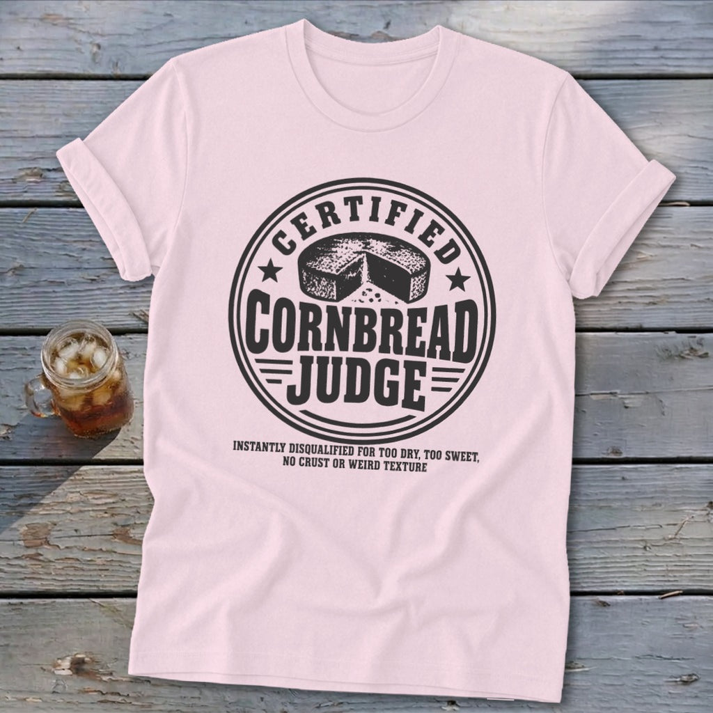 A light pink t-shirt with a black graphic that reads Certified Cornbread Judge on a wooden surface.
