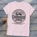 A light pink t-shirt with a black graphic that reads Certified Cornbread Judge on a wooden surface.