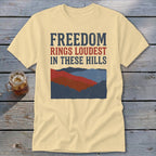 Cream t-shirt featuring distressed blue and red text reading Freedom Rings Loudest in These Hills above a mountain graphic.