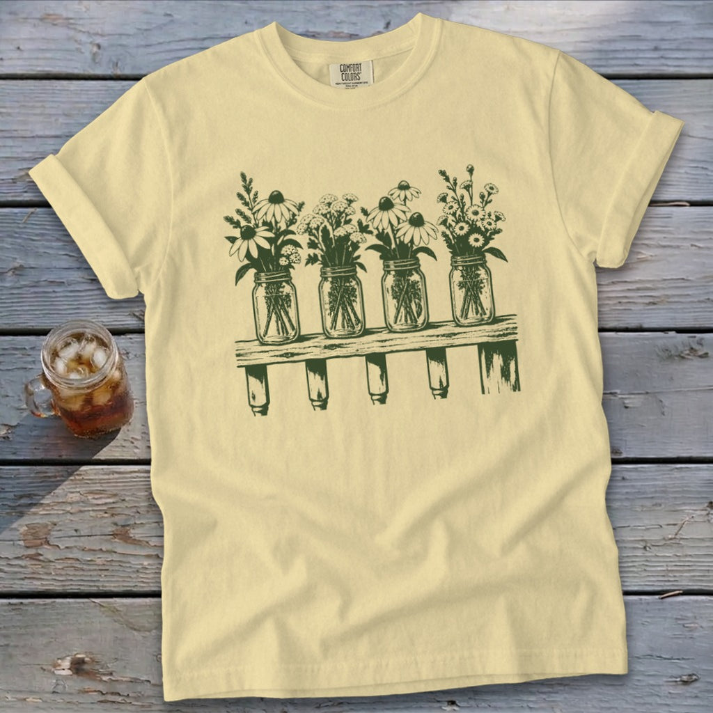 Light yellow t-shirt with a green graphic of four mason jars filled with wildflowers on a shelf.