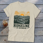 Cream t-shirt with a graphic of mountains at sunrise and the text "RIDGELINE AT DAYBREAK".
