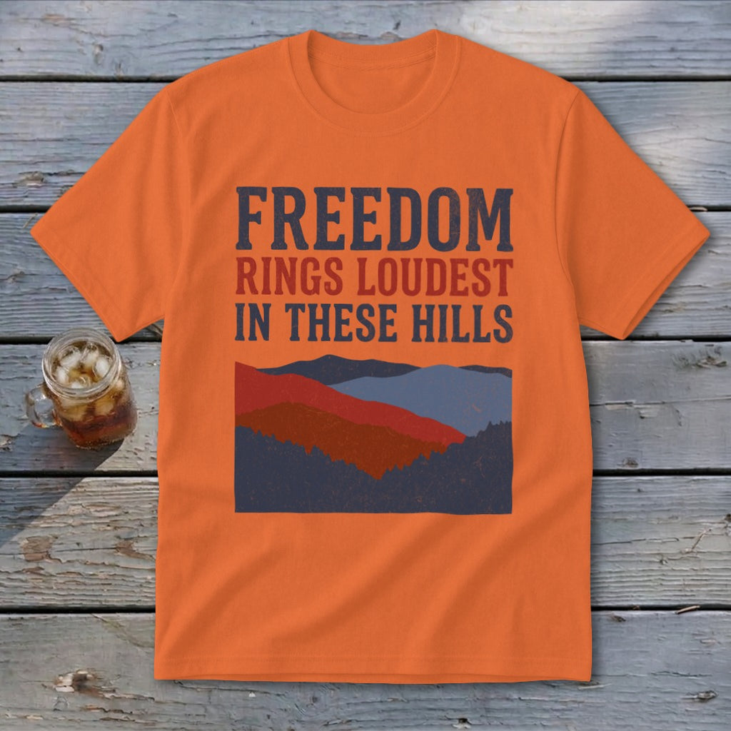 An orange t-shirt featuring the text "FREEDOM RINGS LOUDEST IN THESE HILLS" above layered mountain graphics.