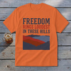 An orange t-shirt featuring the text "FREEDOM RINGS LOUDEST IN THESE HILLS" above layered mountain graphics.
