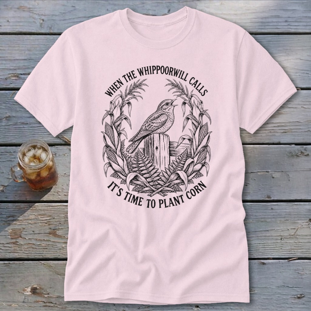 A light pink t-shirt with a vintage-style black illustration of a bird and corn stalks, featuring the text "When the whippoorwill calls it's time to plant corn".