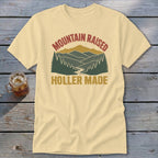 A light yellow t-shirt features a vintage graphic with mountains, pine trees, and the text MOUNTAIN RAISED HOLLER MADE.