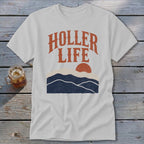 Light gray t-shirt with the text HOLLER LIFE above a sunset landscape graphic on wood background.