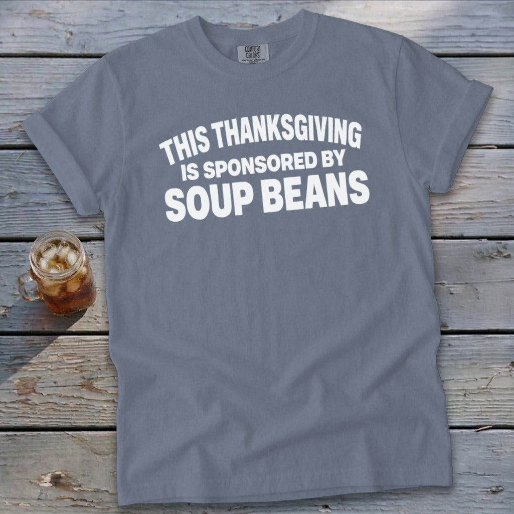 A gray t-shirt with white text reading "This Thanksgiving is sponsored by soup beans" rests on a wooden surface.