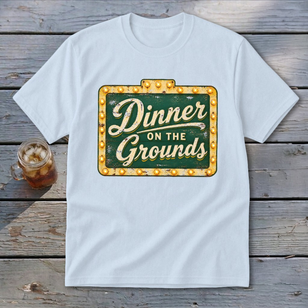 A white crewneck t-shirt featuring a green vintage-style sign with the text "Dinner on the Grounds."