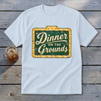 A white crewneck t-shirt featuring a green vintage-style sign with the text "Dinner on the Grounds."