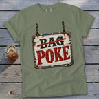 Sage green t-shirt with a graphic of a wooden sign reading Bag Poke, lying on a rustic wooden background.