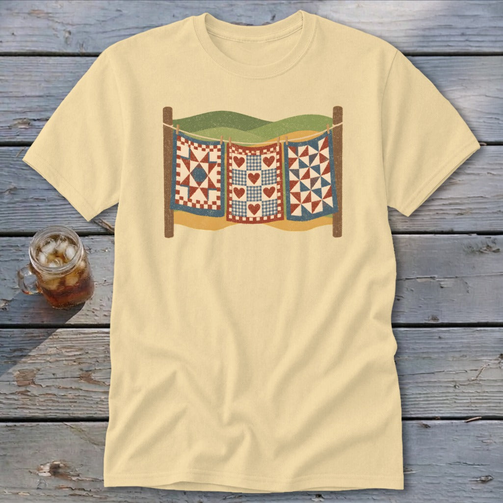 A cream-colored t-shirt with a graphic of three patchwork quilts hanging on a clothesline outdoors.