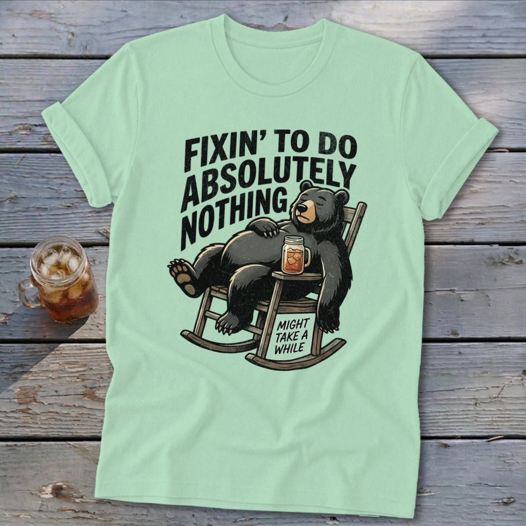 Light green t-shirt with a graphic of a bear lounging in a rocking chair holding a drink, with the text "Fixin' to do absolutely nothing. Might take a while."