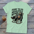 Light green t-shirt with a graphic of a bear lounging in a rocking chair holding a drink, with the text "Fixin' to do absolutely nothing. Might take a while."