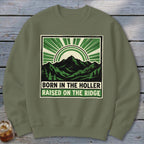 An olive green crewneck sweatshirt featuring a graphic print of mountains, sun rays, and the text Born in the Holler, Raised on the Ridge.