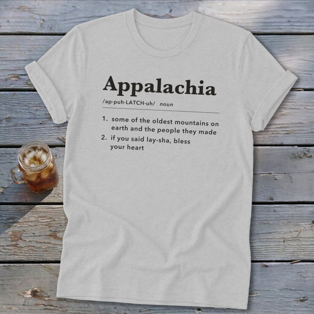 A light gray crewneck t-shirt featuring dictionary-style text defining Appalachia lies flat on a rustic wooden table next to a glass of iced tea.