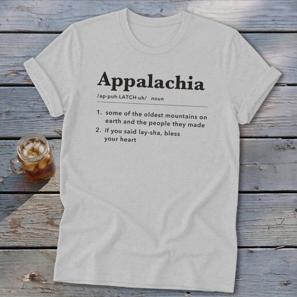 A light gray crewneck t-shirt featuring dictionary-style text defining Appalachia lies flat on a rustic wooden table next to a glass of iced tea.