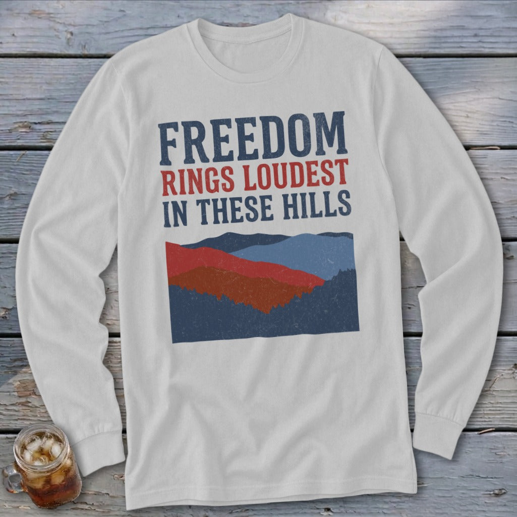 Light gray long-sleeved t-shirt featuring distressed blue and red text reading Freedom Rings Loudest in These Hills above a mountain landscape graphic.