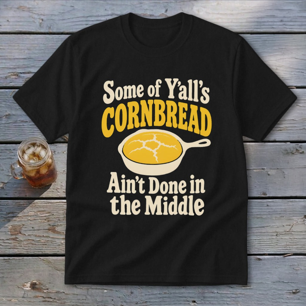 A black t-shirt printed with the text Some of Y'all's Cornbread Ain't Done in the Middle and a skillet graphic.
