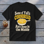 A black t-shirt printed with the text Some of Y'all's Cornbread Ain't Done in the Middle and a skillet graphic.