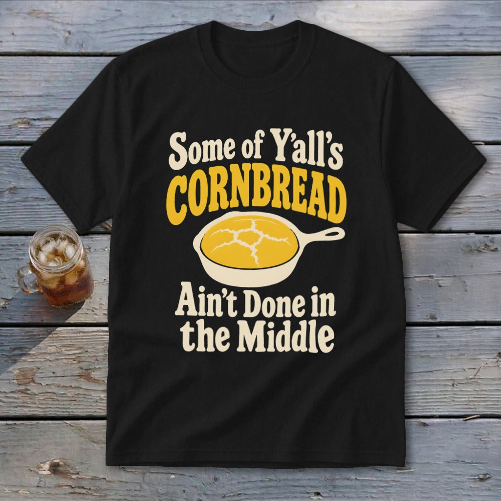A black t-shirt printed with the text Some of Y'all's Cornbread Ain't Done in the Middle and a skillet graphic.