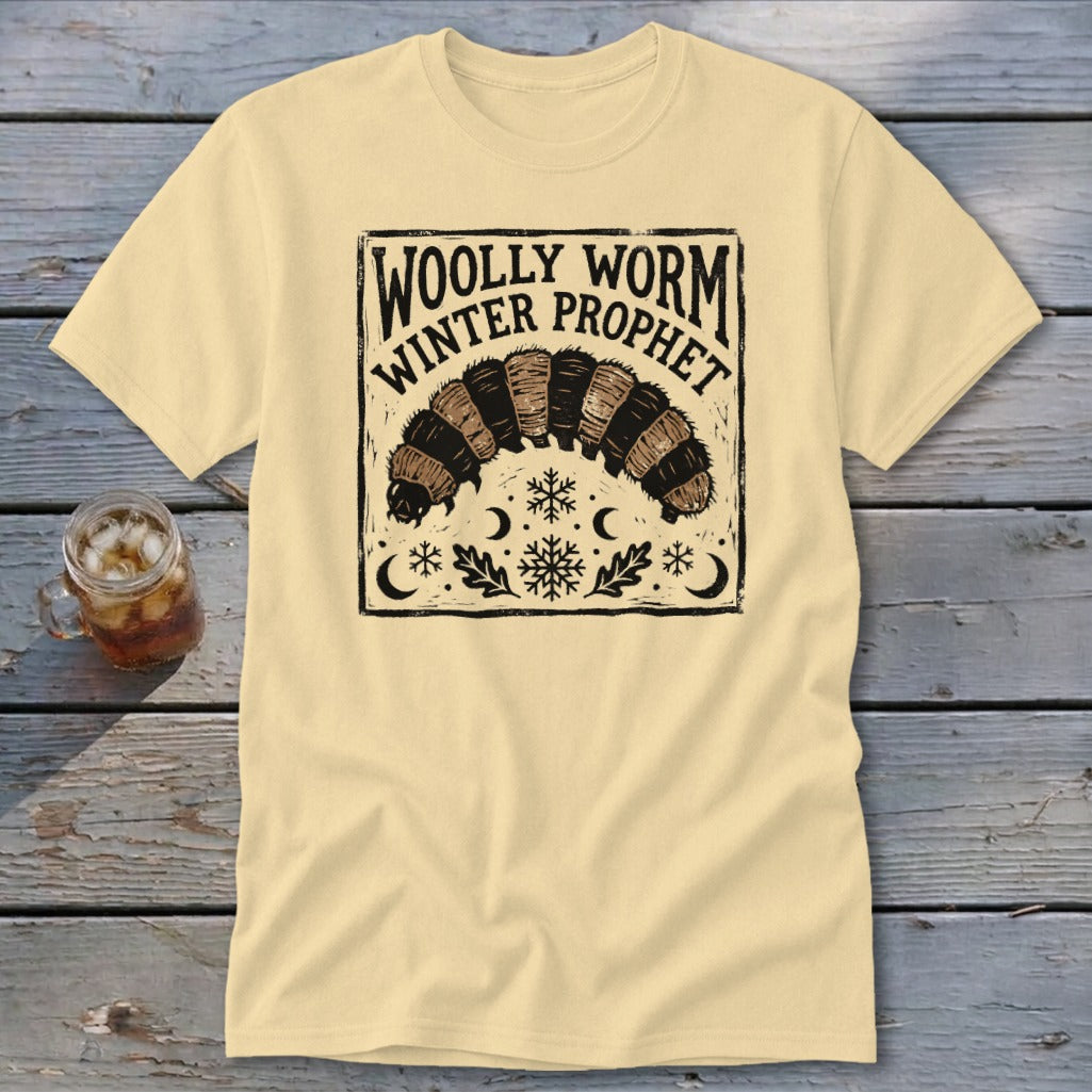 A cream-colored t-shirt with a vintage black graphic of a woolly worm caterpillar and text reading WOLLY WORM WINTER PROPHET.