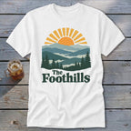 White t-shirt with a graphic of mountains, sun, and the text "The Foothills".