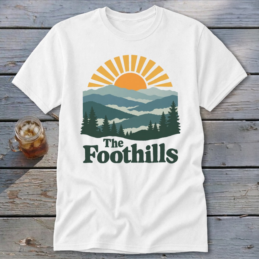 White t-shirt with a graphic of mountains, sun, and the text "The Foothills".