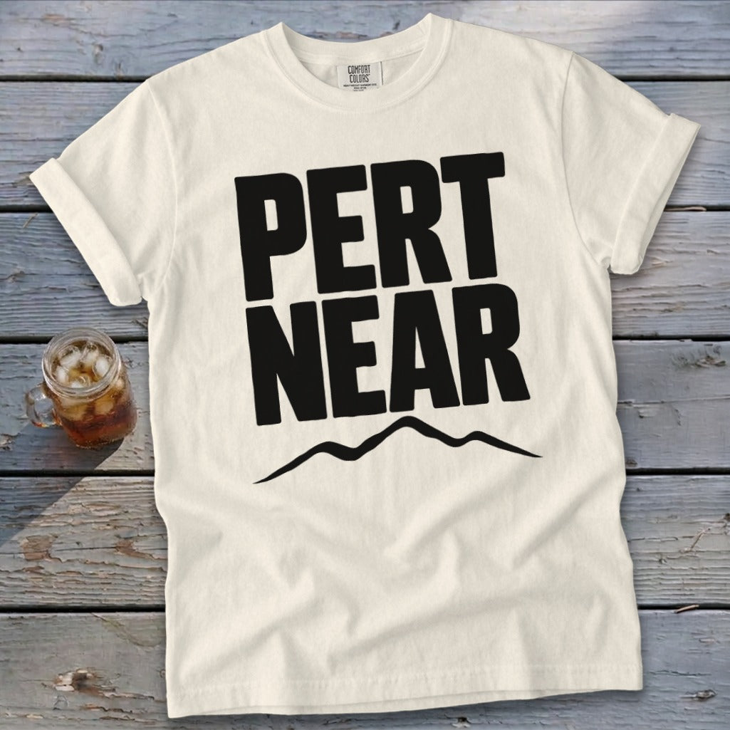 A white t-shirt with black text reading PERT NEAR above a mountain range graphic, laid on a wooden surface next to a glass of iced tea.