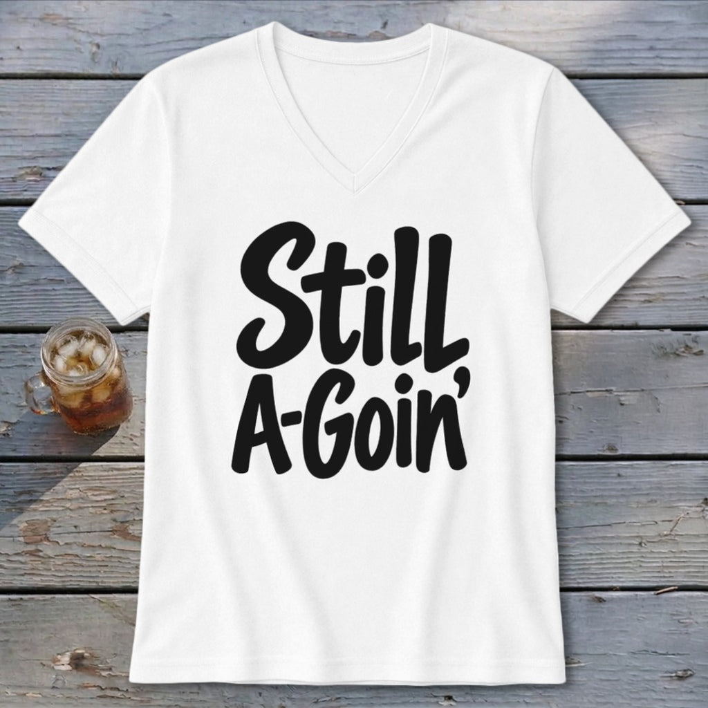 A white V-neck t-shirt with the black text "Still A-Goin'" printed on the front, displayed on a wooden surface beside a glass of iced tea.