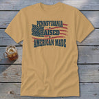 A tan t-shirt with distressed text reading "PENNSYLVANIA RAISED AMERICAN MADE" and a distressed American flag graphic.