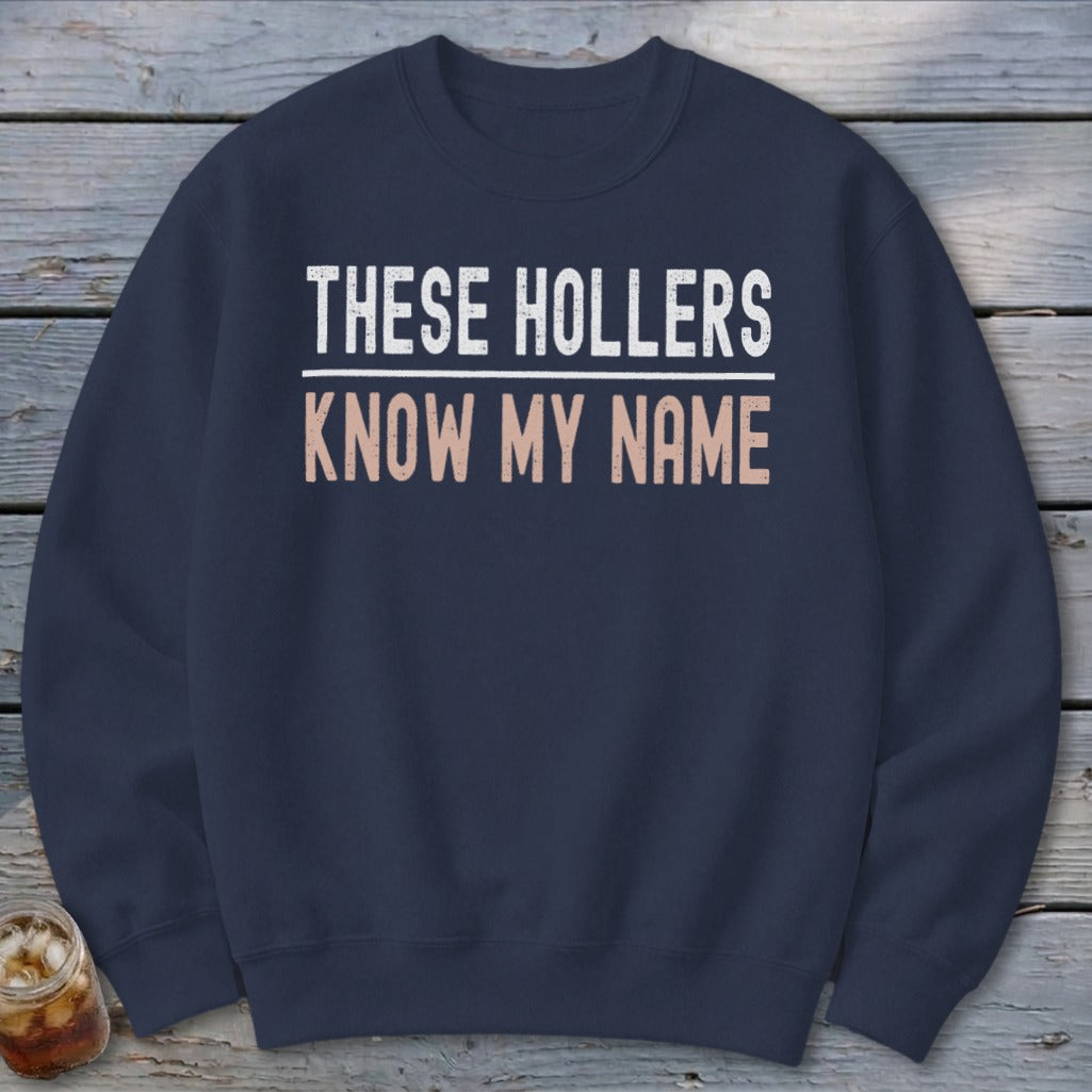 Navy sweatshirt with white text reading "THESE HOLLERS" and tan text reading "KNOW MY NAME".
