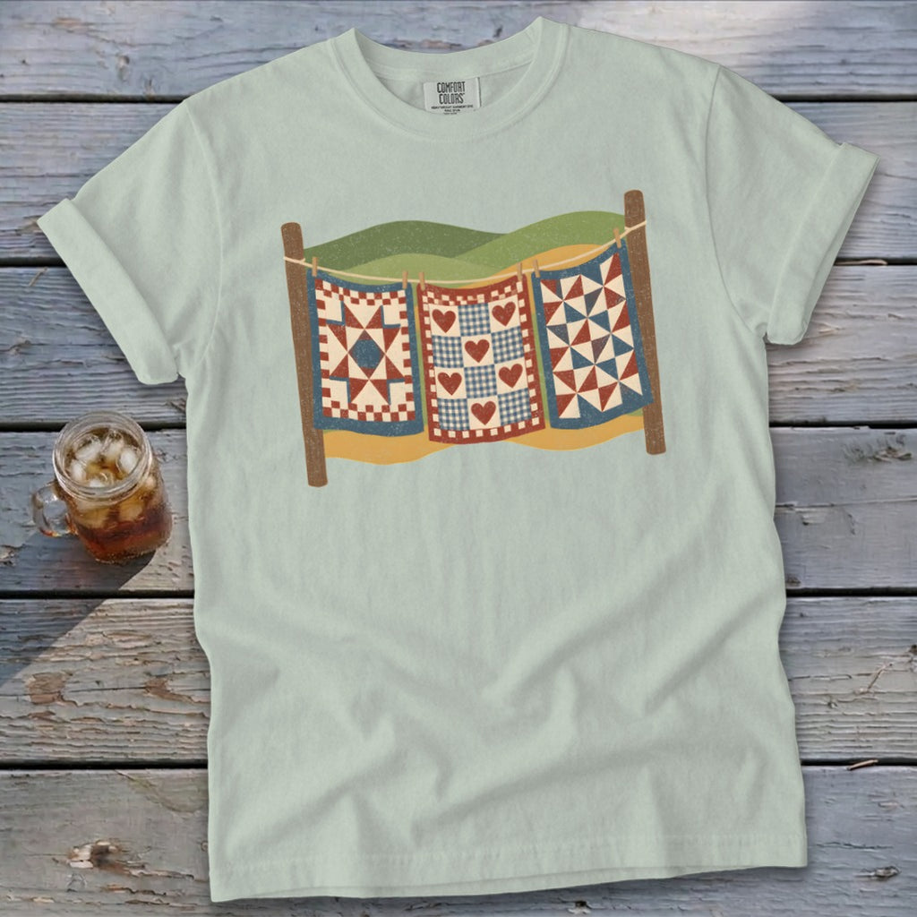 Light green t-shirt featuring a graphic of three patchwork quilts hanging on a clothesline outdoors.